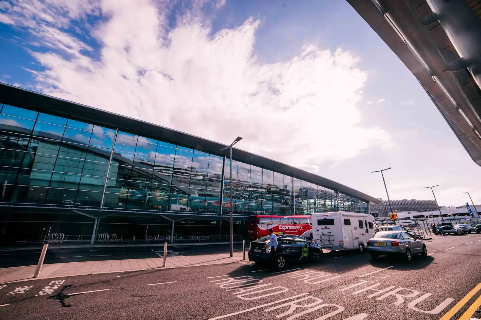 Glass airport terminal with dark cars on the arrivals road under a bright sky