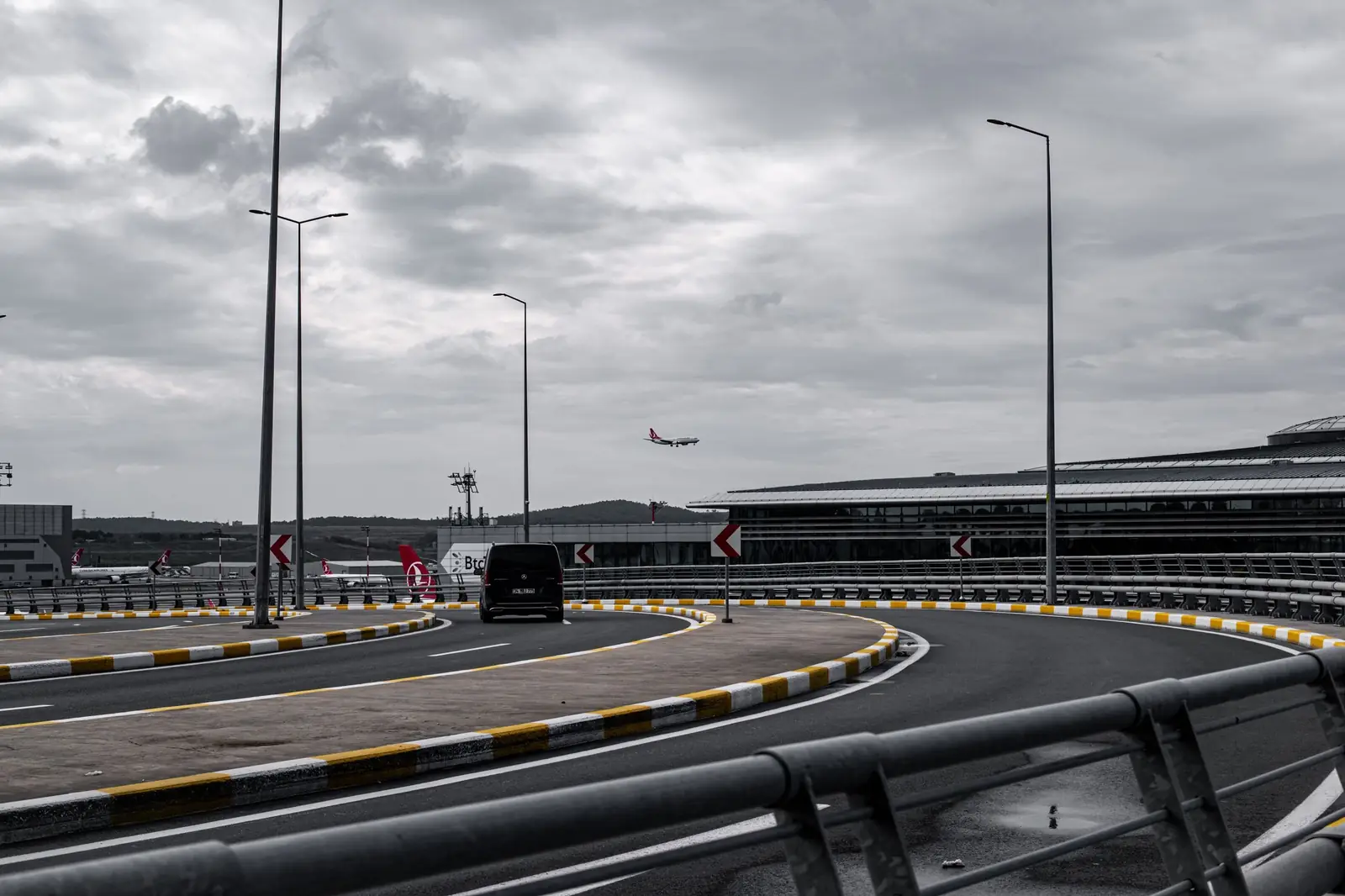 Curved airport access road with a black van and a plane landing in the grey sky
