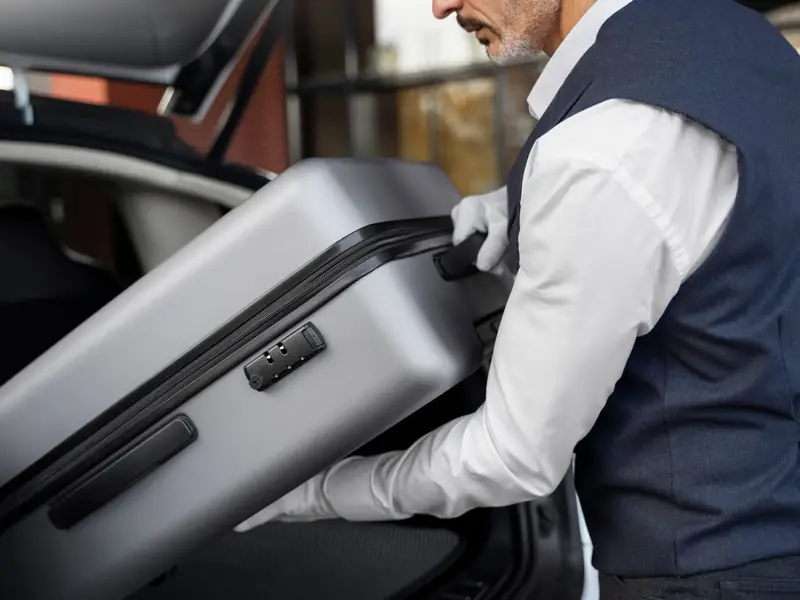 Chauffeur in white gloves lifting a grey suitcase into the boot of a car