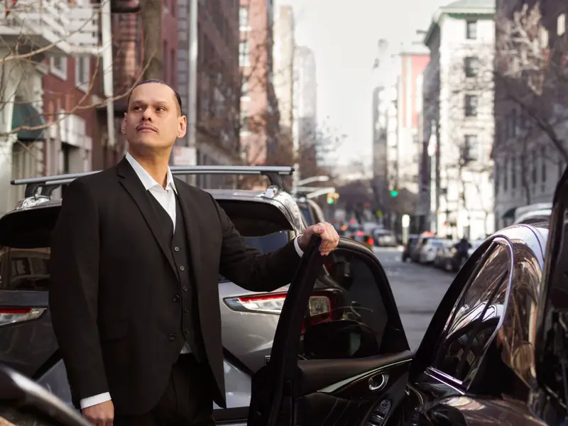 Chauffeur in a black suit holding the door of a black car open on a city street