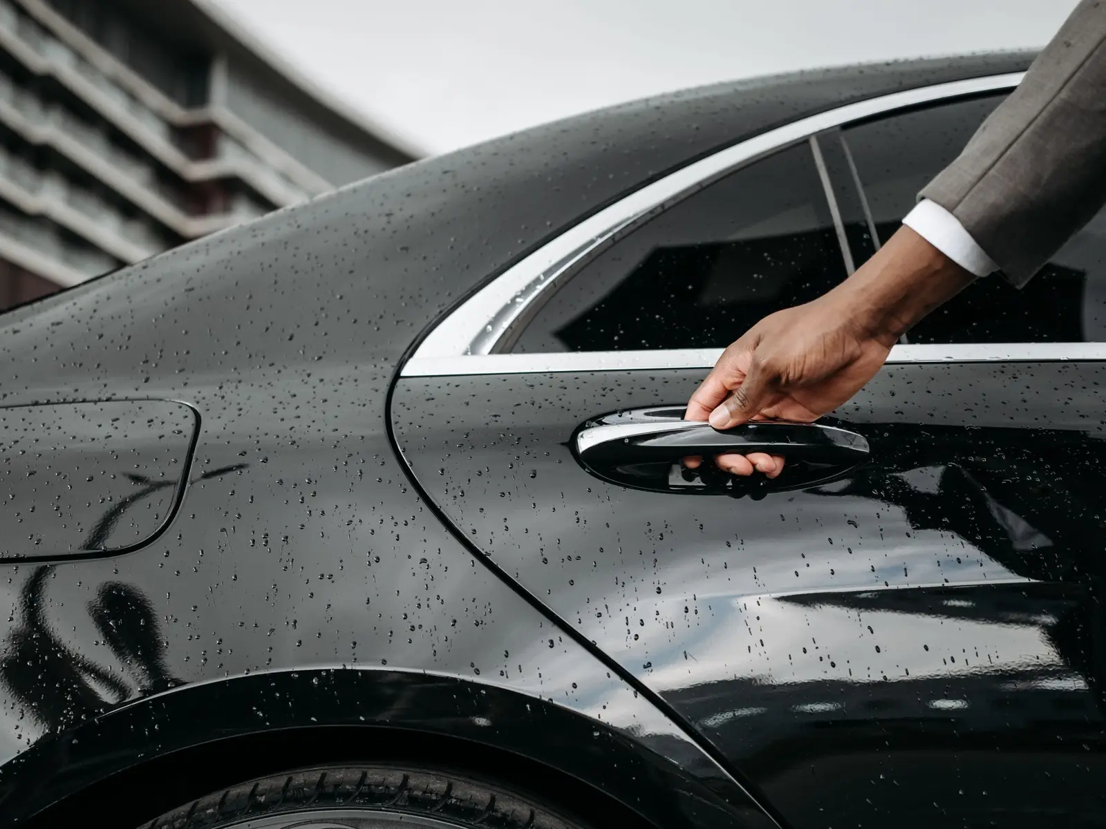 Chauffeur's hand on the door handle of a black sedan with raindrops on the paint