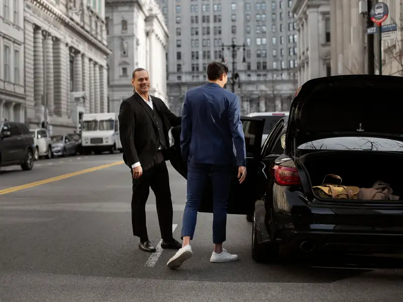 Chauffeur opening the rear door of a black car for a client on a New York street, bags in the open boot