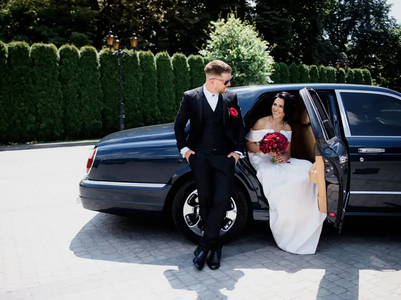 Groom leaning on a dark classic car while the bride sits in the back seat holding red flowers
