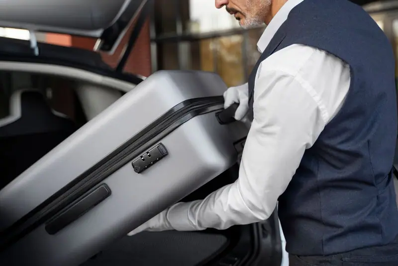 Chauffeur in white gloves lifting a grey suitcase into the boot of a car