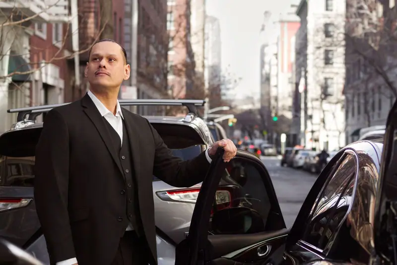 Chauffeur in a black suit holding the door of a black car open on a city street