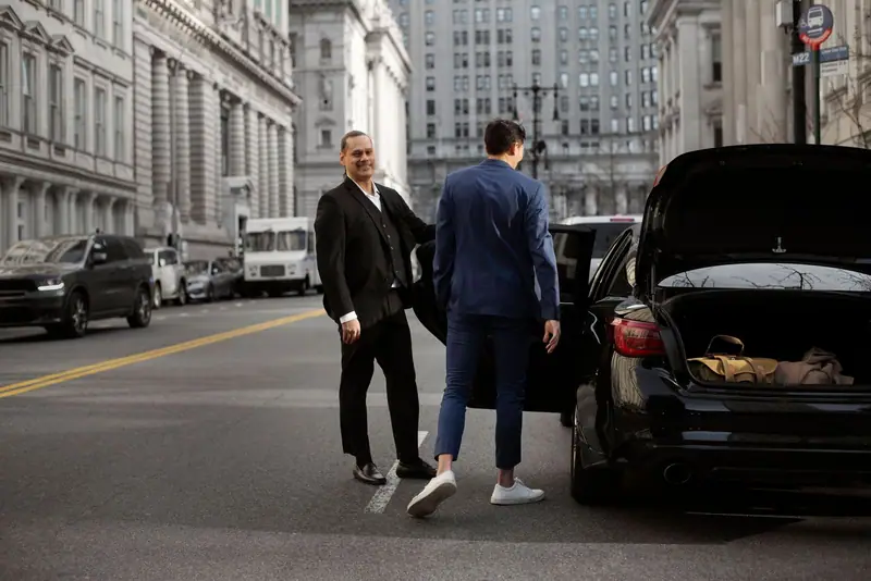 Chauffeur opening the rear door of a black car for a client on a New York street, bags in the open boot