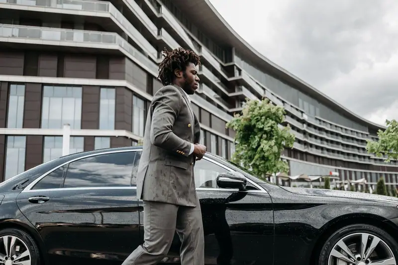 Businessman in a grey suit walking past a black Mercedes-Benz sedan in front of a hotel