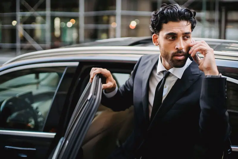 Businessman in a dark suit taking a phone call as he steps out of a black car