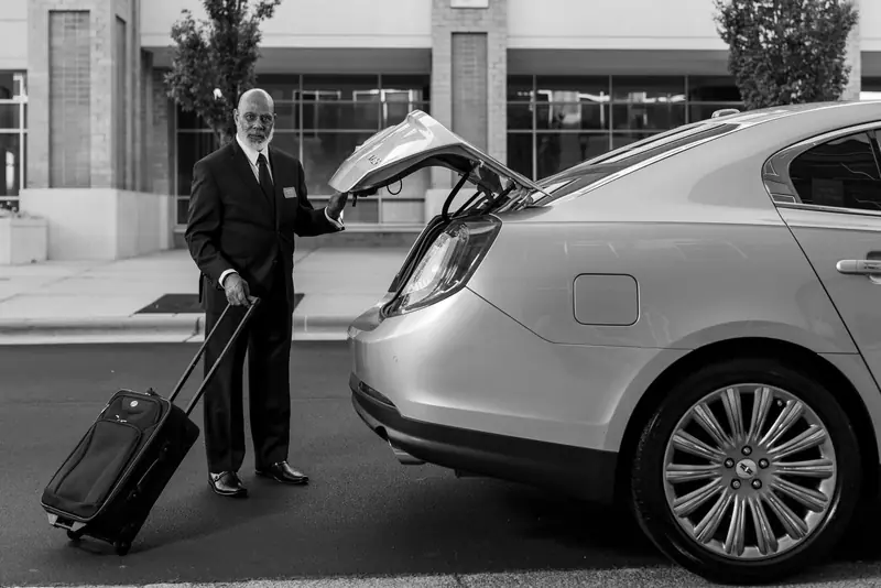 Chauffeur in a black suit loading a suitcase into the boot of a sedan, in black and white