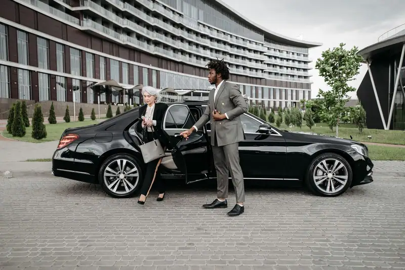 Chauffeur in a grey suit helping an older passenger out of a black Mercedes-Benz S-Class at a hotel