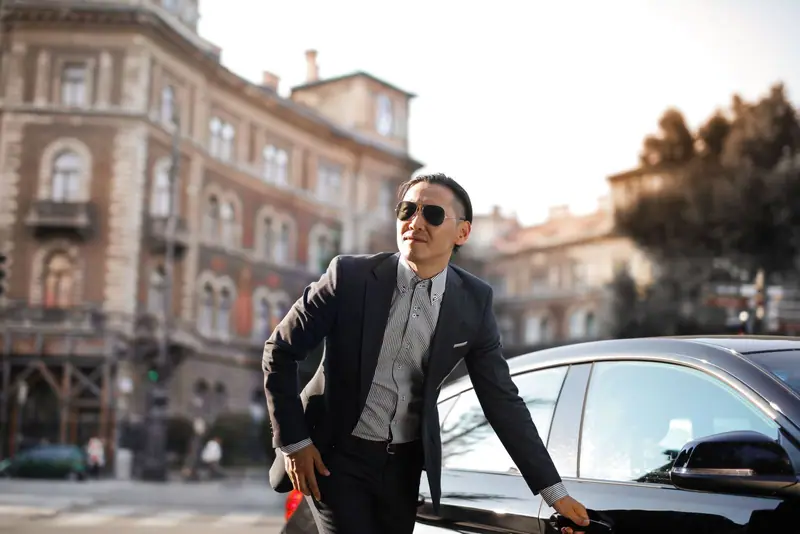 Man in sunglasses and a dark jacket leaning on a black car on a city street