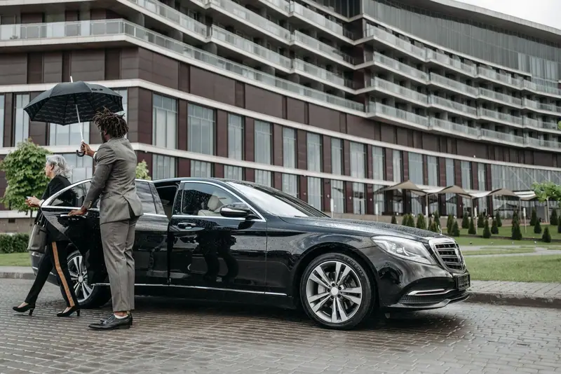 Chauffeur holding an umbrella while a passenger gets out of a black Mercedes-Benz S-Class outside a hotel