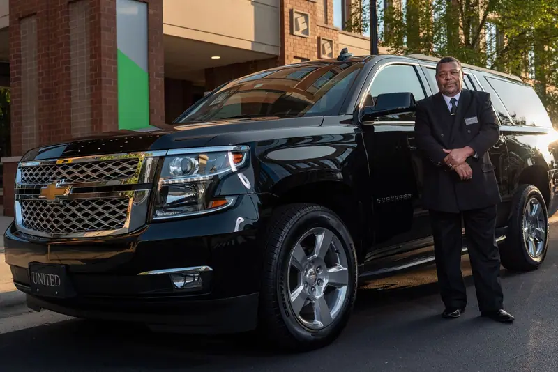 Chauffeur in a dark suit standing beside a black Chevrolet Suburban outside a hotel