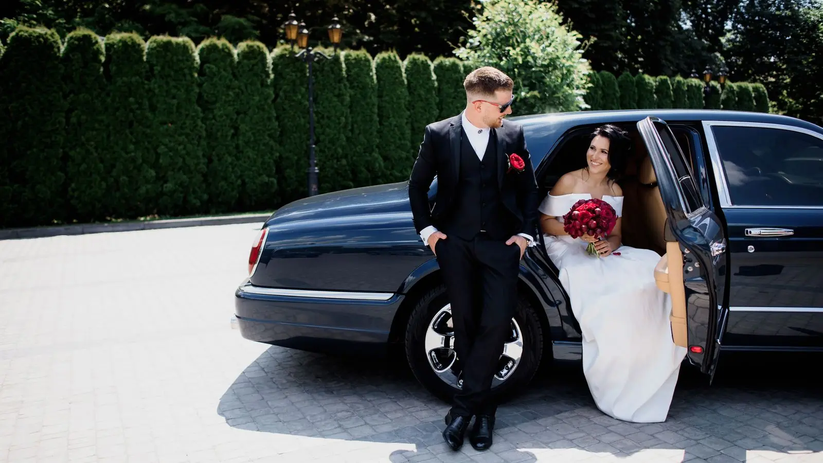 Groom leaning on a dark classic car while the bride sits in the back seat holding red flowers