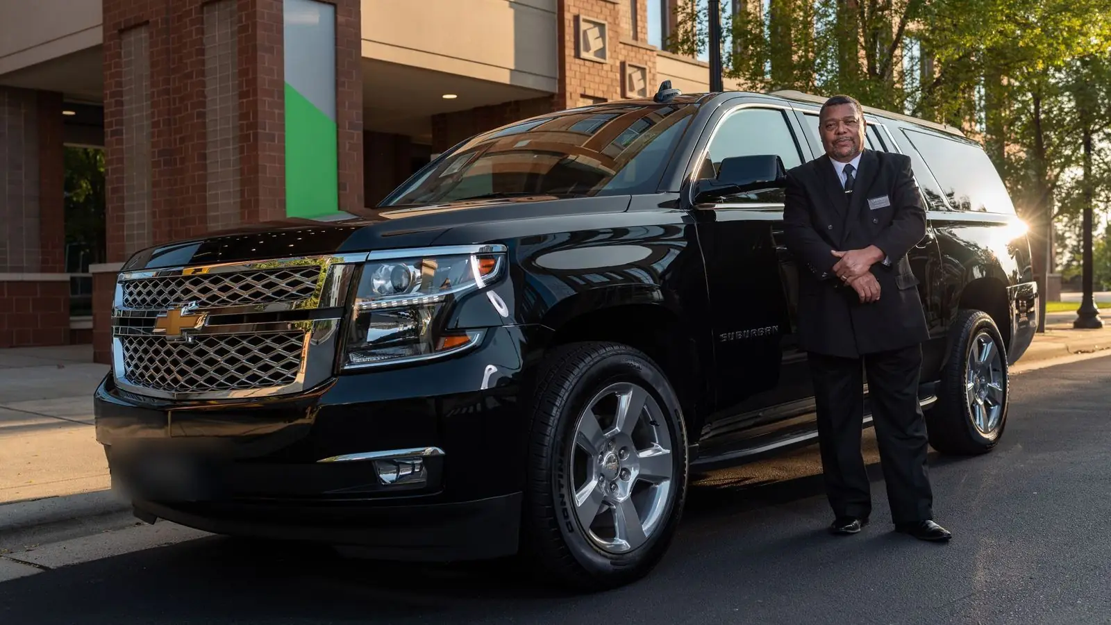 Chauffeur in a dark suit standing beside a black Chevrolet Suburban outside a hotel