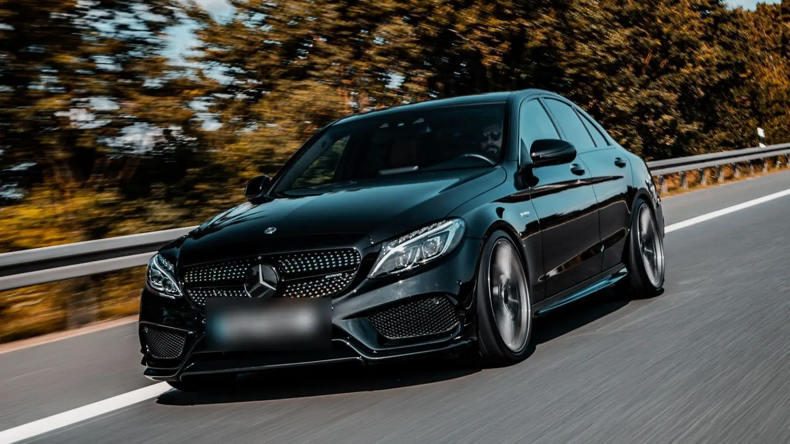 Black Mercedes-Benz C-Class driving along a road lined with autumn trees